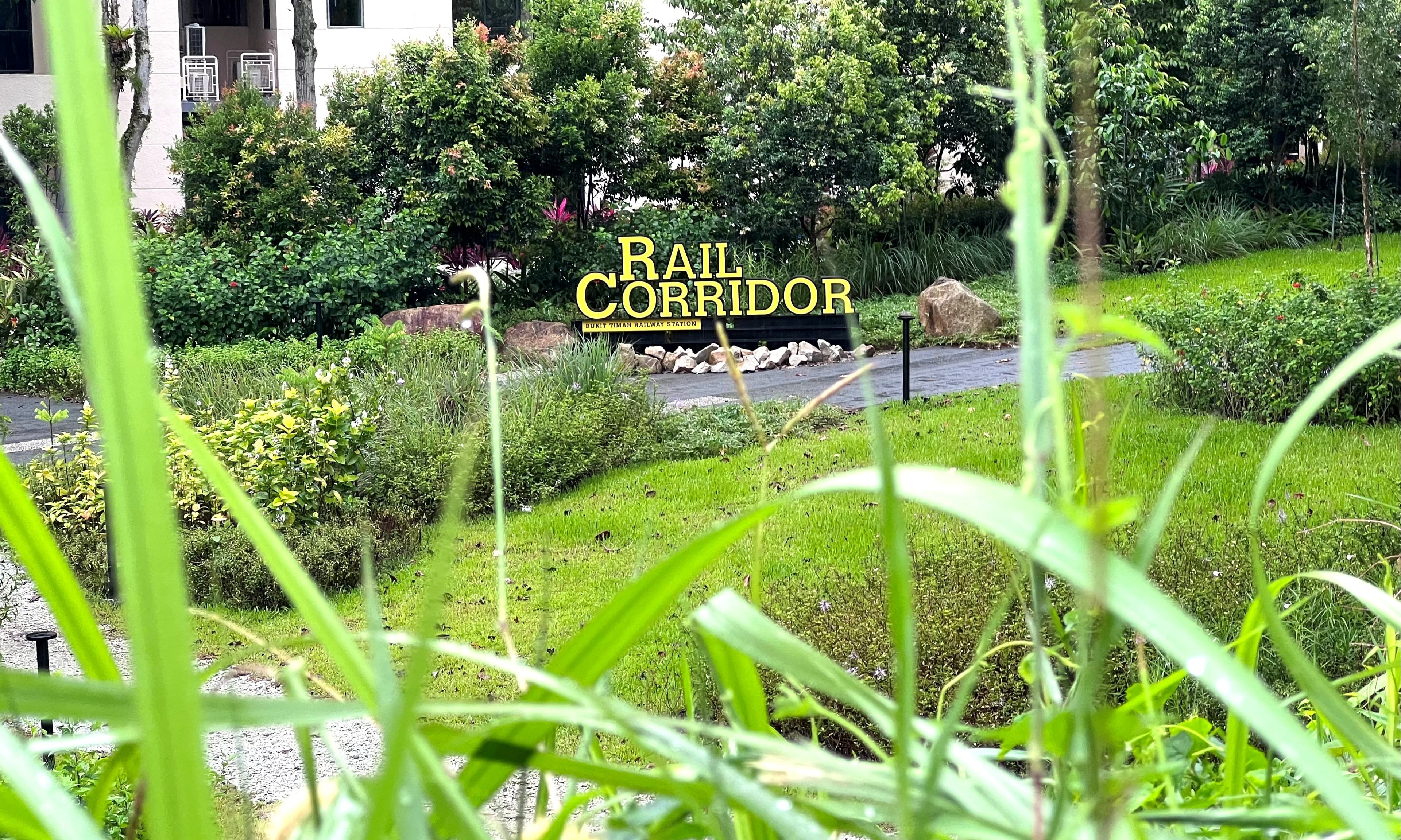 A sign reading "Rail Corridor" stands in a lush green park with trees and grass, with a building in the background.